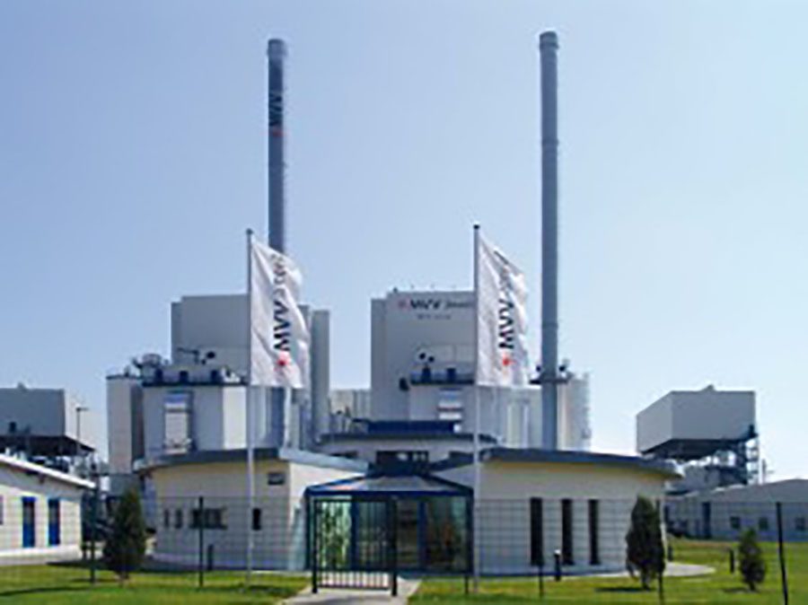 Kraftwerksgebäude mit zwei hohen Schornsteinen und Firmenflaggen vor Eingang.