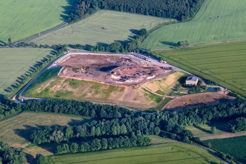 Luftbild einer von Feldern und Wald umgebenen Deponiefläche mit Erdaufschüttungen, Fahrzeugen und einem kleinen Betriebsgebäude.