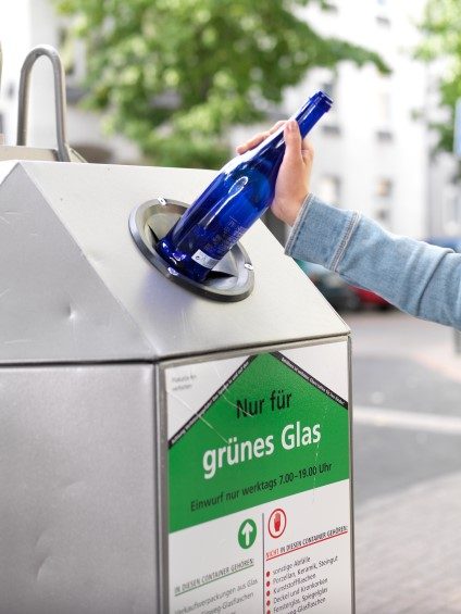 Hand wirft blaue Glasflasche in Container für grünes Glas ein.